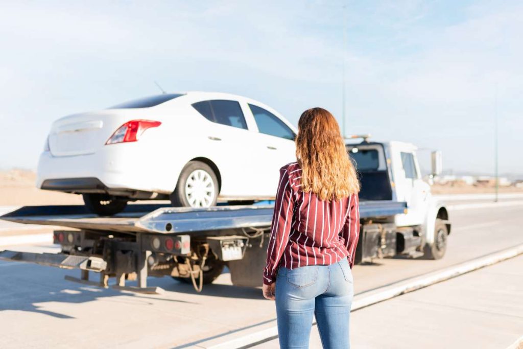 Rear view of a blond young woman watching her car being towed on a tow truck on the side of the highway